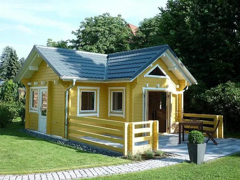 Ein kleines gelbes Holzhaus mit weißer Verkleidung, einem grauen Ziegeldach und einer Veranda. Das Haus steht auf einem grasbewachsenen Rasen, mit Bäumen im Hintergrund und einem gepflasterten Weg, der zum Eingang führt.