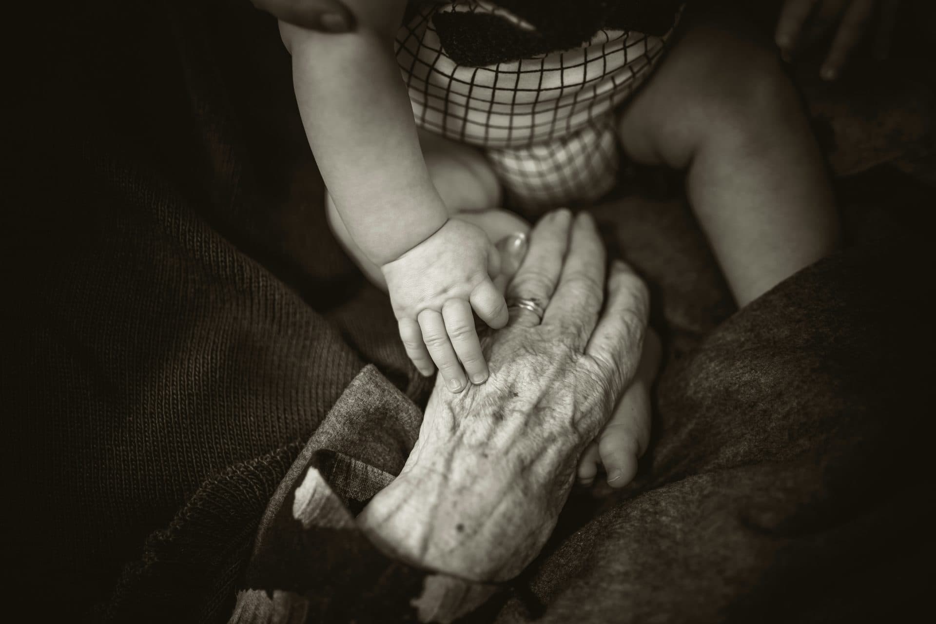 ein sepiafarbenes bild zeigt ein baby, das sanft die faltige hand einer älteren person berührt und so die verbindung zwischen den generationen symbolisiert. das baby trägt ein kariertes outfit, und die szene wirkt warm und intim.